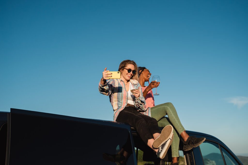Multiracial friends having fun doing video call celebrating with wine while sitting on top of camper